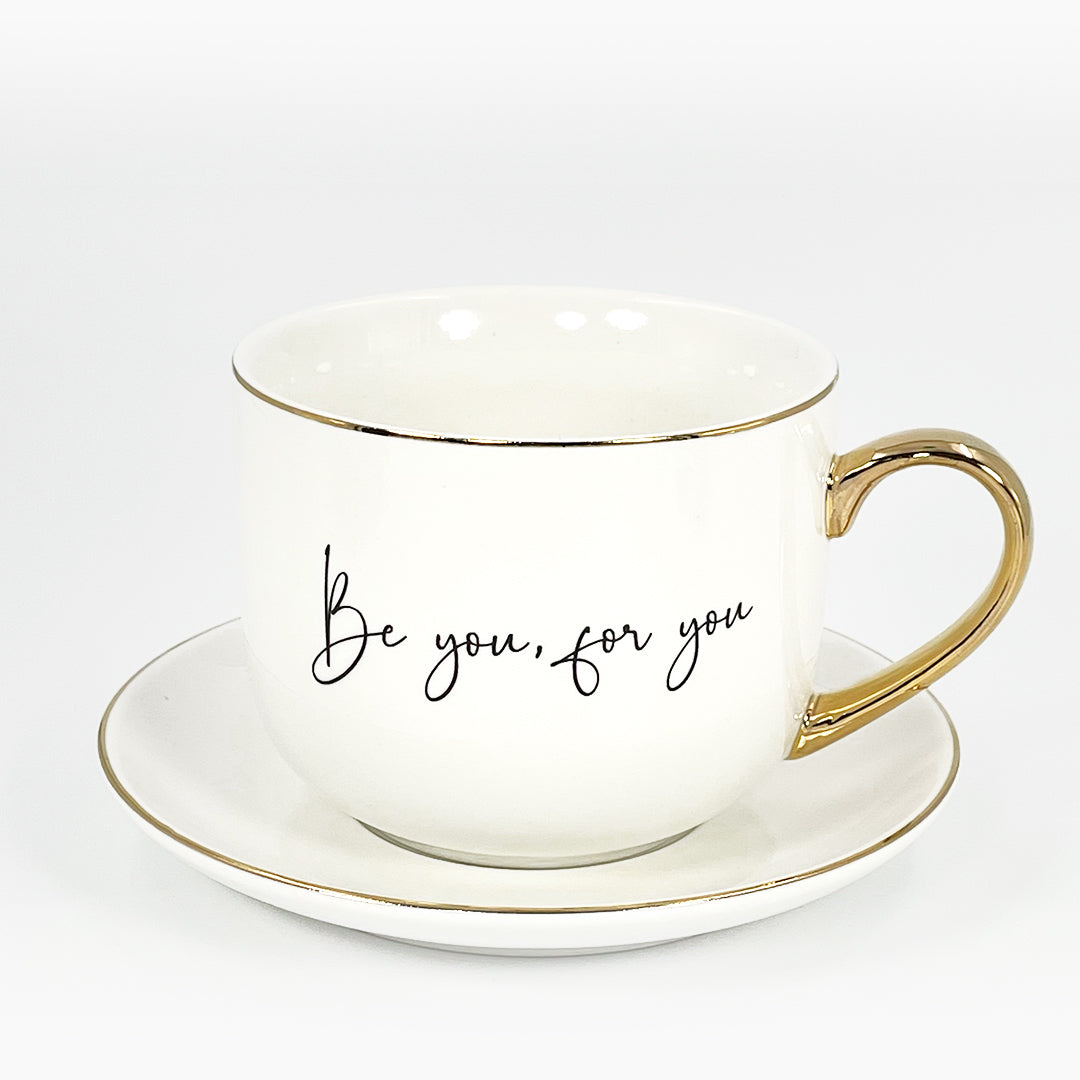 Leftt side of a white ceramic cup and saucer with 24k gold rim and handle and with the text "Be you, For you" written on it.