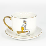 The right side of a white ceramic cup and saucer with 24k gold rim and handle with an image of a girl sitting on it.